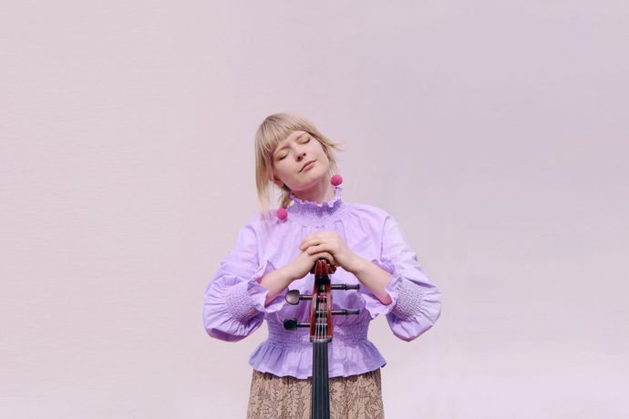 Woman in a purple blouse gently holds a cello, eyes closed, against a soft pastel background.
