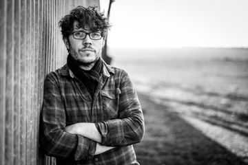 Black and white photo of a person leaning against a wooden wall with arms crossed, wearing a plaid shirt and scarf, with an open landscape in the background.