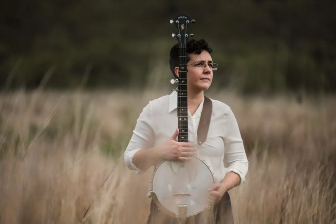 Person standing in tall grass holding a banjo vertically in front of them, wearing glasses and a white shirt, looking to the right.