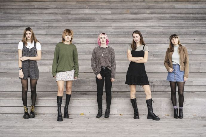 Five young women with diverse styles stand in a line against a wooden wall, wearing skirts, dresses, and boots.