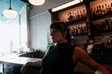 A person sits casually at a bar counter in a dimly lit space with shelves of bottles behind them and large windows to the left letting in natural light. Provide your feedback on BizChat