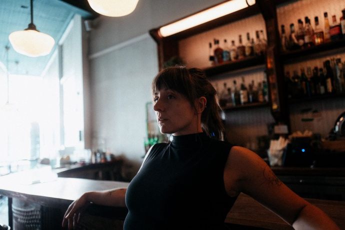 A person sits casually at a bar counter in a dimly lit space with shelves of bottles behind them and large windows to the left letting in natural light. Provide your feedback on BizChat