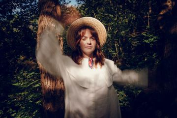 A person wearing a straw hat and white blouse stands in a sunlit forest, with blurred arms suggesting movement.