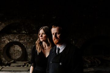 A man and woman stand in a dimly lit, rustic space with brick walls. The woman looks left, the man faces forward, both in dark clothing.