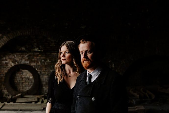 A man and woman stand in a dimly lit, rustic space with brick walls. The woman looks left, the man faces forward, both in dark clothing.