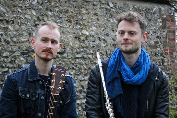 Two men sitting outdoors against a stone wall, one holding a guitar and the other a flute, both wearing jackets and scarves.
