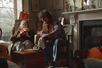 Two people enjoy tea in a cozy living room, surrounded by musical instruments and soft light from a large window.