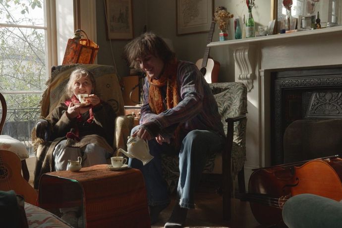 Two people enjoy tea in a cozy living room, surrounded by musical instruments and soft light from a large window.