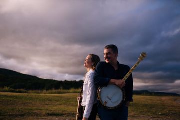 A man and woman stand outside with musical instruments, the man holding a banjo. They are smiling, with a cloudy sky and open field behind them.