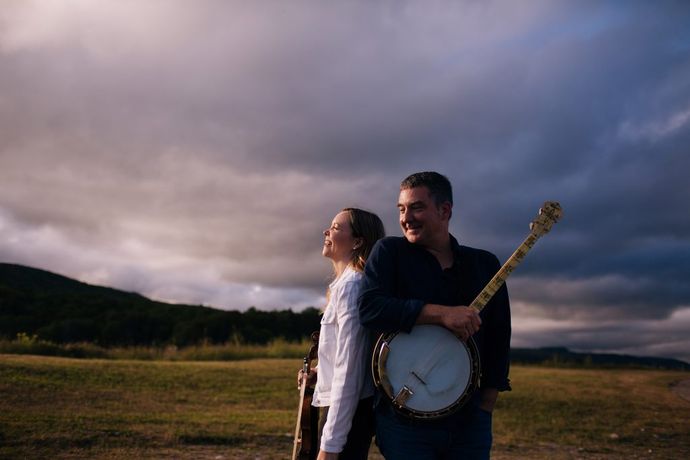 A man and woman stand outside with musical instruments, the man holding a banjo. They are smiling, with a cloudy sky and open field behind them.