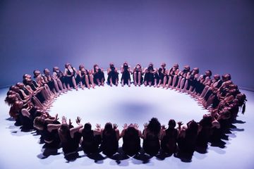 A group of people in dark clothing sit in a large circle under purple lighting, facing inward with their hands raised and outstretched.