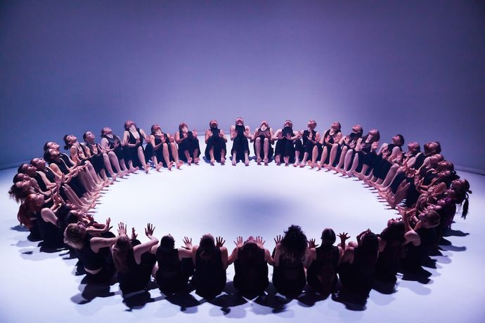 A group of people in dark clothing sit in a large circle under purple lighting, facing inward with their hands raised and outstretched.
