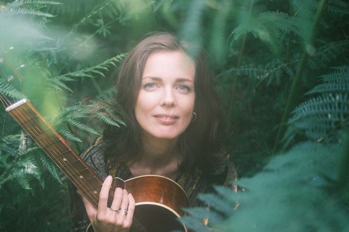 A person with long hair holds a guitar, surrounded by lush green foliage, gazing upward with a serene expression.