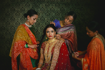 A bride in a red and gold sari is being adorned with jewelry by three women against a patterned background.