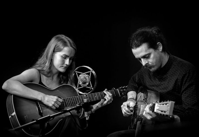 Black and white image of Kelvin Davies & Chloe Turner playing acoustic guitars with focus, a vintage microphone between them.