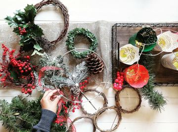 A person arranges festive wreaths, pinecones, berries, and colorful paper lanterns on a table, creating a holiday decoration display.