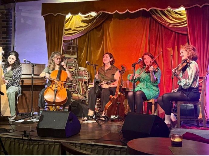 A small acoustic ensemble performs on an intimate stage, seated with string instruments in front of microphones and warm-colored curtains.