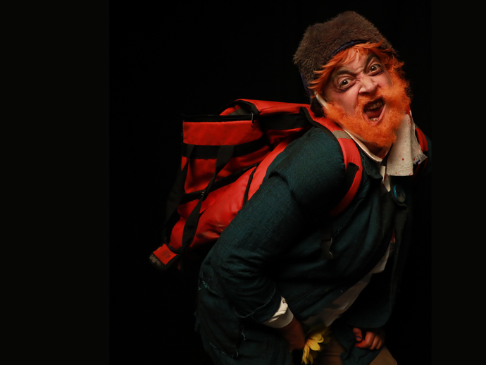 Person in colorful costume and orange beard, wearing a big red backpack, poses animatedly against a black background.