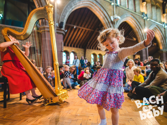 A young girl dances joyfully in a church hall with people seated around her, and a harpist performing nearby.