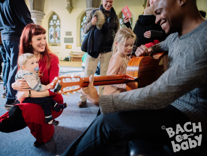 A man plays guitar while children and adults gather around, engaging with the music in a lively, social setting.