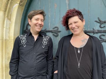 Two people standing in front of a large teal wooden door with ornate black metal hinges. Both wear black clothing; one has a patterned shirt, and the other has layered necklaces and a visible tattoo on the chest.