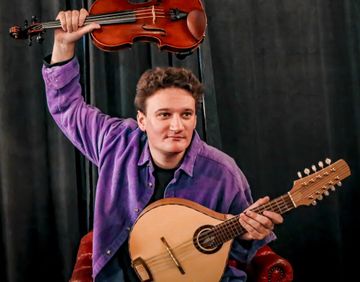 A person in a purple shirt holds a violin above their head and a stringed instrument in the other hand, sitting on a red chair against a dark background.