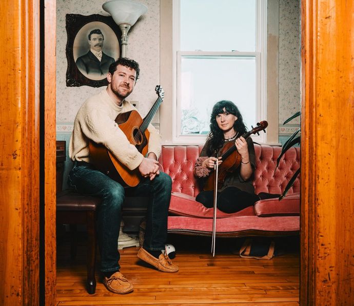 Two musicians sit on a pink couch in a cozy room; one holds a guitar, the other a violin. A vintage portrait hangs on the wall behind them.