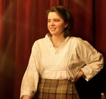 Person wearing white blouse and beige tartan skirt stands with hand on hip looking to side