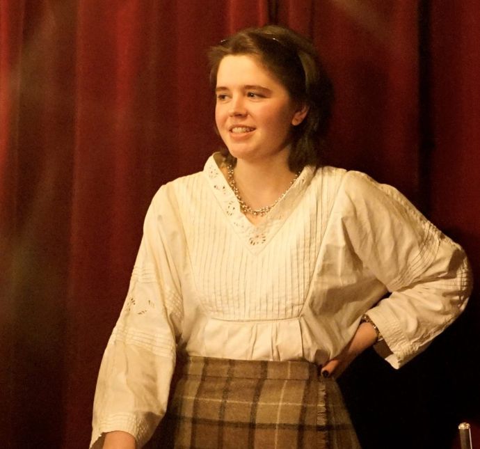 Person wearing white blouse and beige tartan skirt stands with hand on hip looking to side