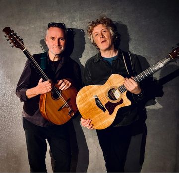 Two people are standing against a textured gray wall, each holding an acoustic guitar with warm wooden tones and visible strings.