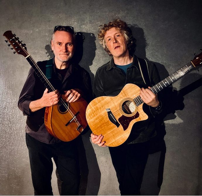 Two people are standing against a textured gray wall, each holding an acoustic guitar with warm wooden tones and visible strings.