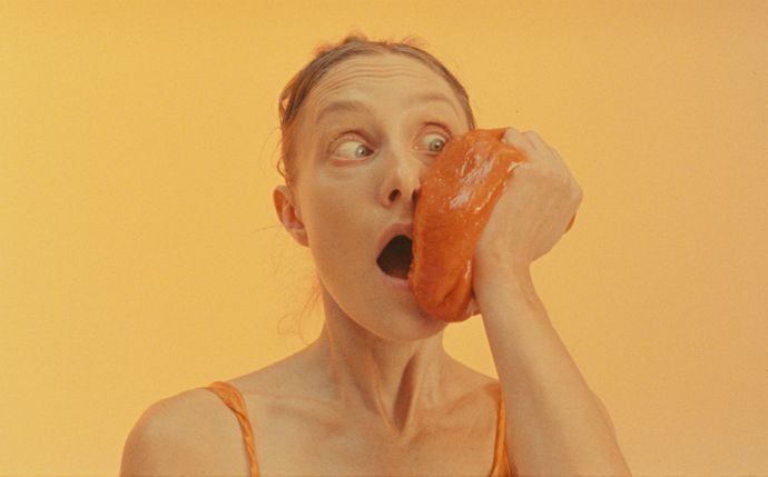 Yellow background with person looking shocked holding orange shiny squishy object to cheek