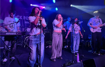Band performing on stage with colorful lights, wearing matching shirts with peace symbols. Keyboardist, singers, and guitarist are visible.