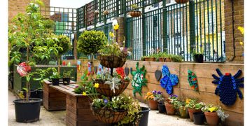 Urban garden with colorful flowers, potted plants, and wall art made from recycled materials against a green fence.
