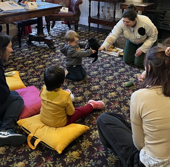 Children sit in a circle as a toddler gently holds a small black animal indoors.