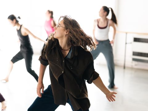 A group of dancers practicing in a studio, each moving with expressive gestures. The focus is on a person with long hair, wearing a brown shirt.