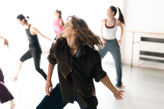 A group of dancers practicing in a studio, each moving with expressive gestures. The focus is on a person with long hair, wearing a brown shirt.