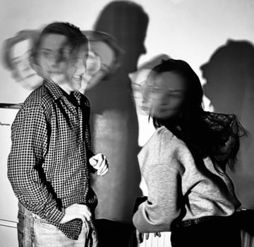 Two people in a room with blurred faces, casting clear shadows on the wall, creating an abstract, ghostly effect. Black and white photo.