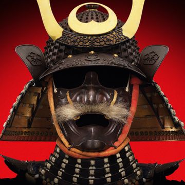 Samurai helmet with ornate gold and black decorations, featured on a red background, displaying intricate metalwork and detailing.