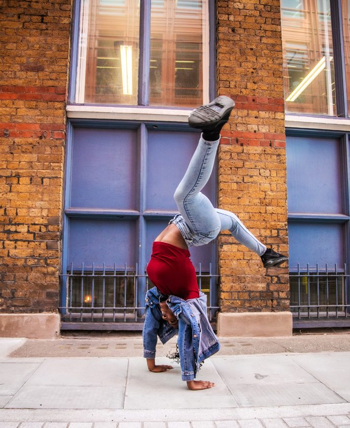 Person in a red shirt and jeans performing a handstand on a city sidewalk, with a brick building in the background.
