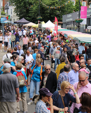 A busy outdoor street fair scene shows crowds of people walking between colourful market stalls lined along a closed-off road.
