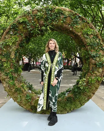 A person standing in front of a large circular sculpture made of moss and plants in an outdoor setting with trees in the background.
