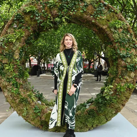 A person standing in front of a large circular sculpture made of moss and plants in an outdoor setting with trees in the background.