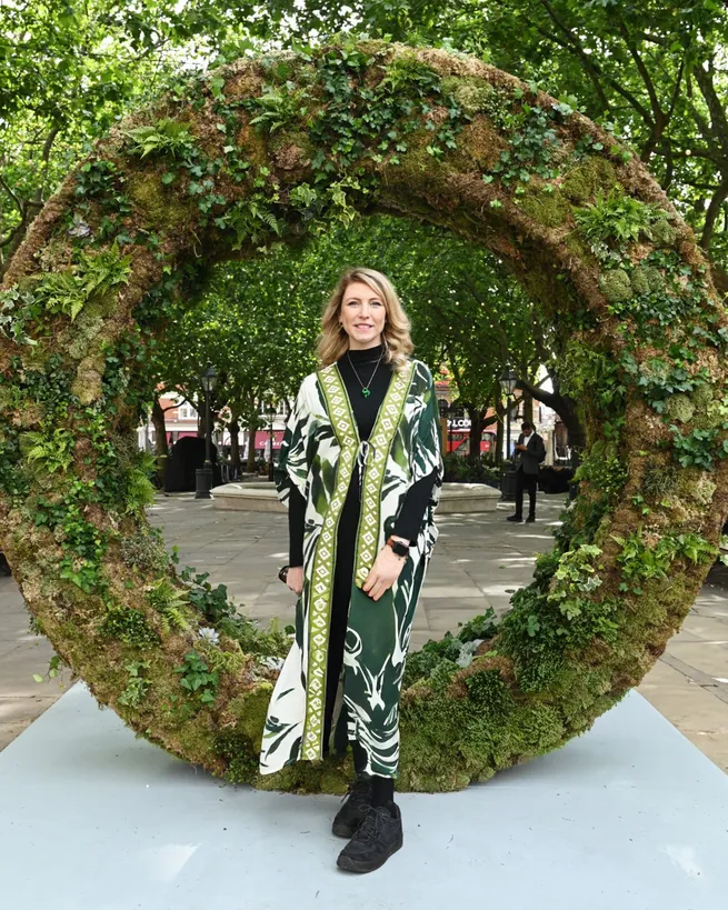 A person standing in front of a large circular sculpture made of moss and plants in an outdoor setting with trees in the background.