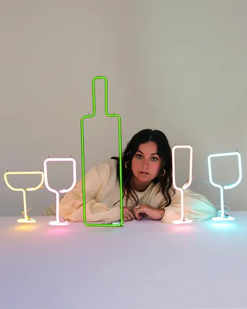 A person leans on a table with colorful neon lights shaped like a bottle and glasses in front of them, against a plain background.