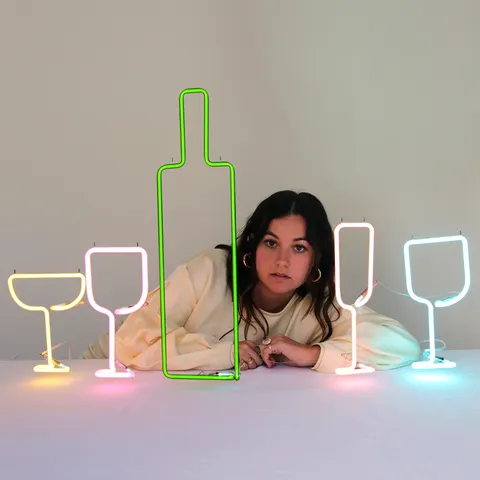A person leans on a table with colorful neon lights shaped like a bottle and glasses in front of them, against a plain background.