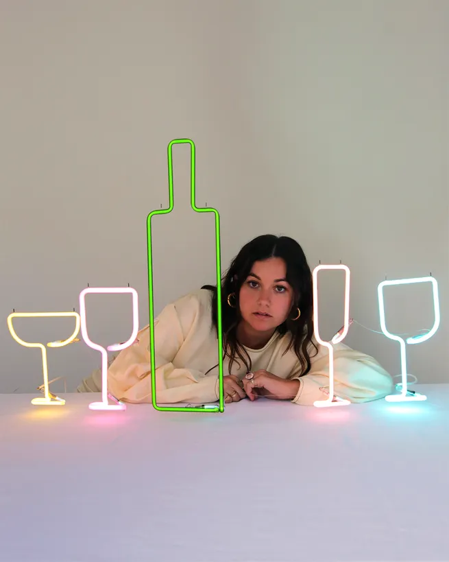 A person leans on a table with colorful neon lights shaped like a bottle and glasses in front of them, against a plain background.