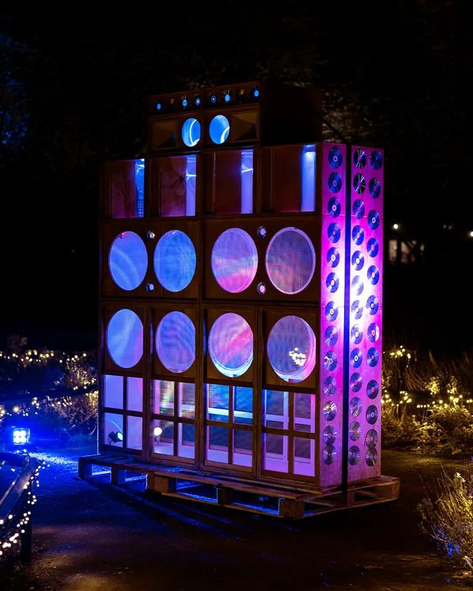 Large illuminated wooden structure with circular and rectangular openings, emitting colorful lights, set outdoors at night.