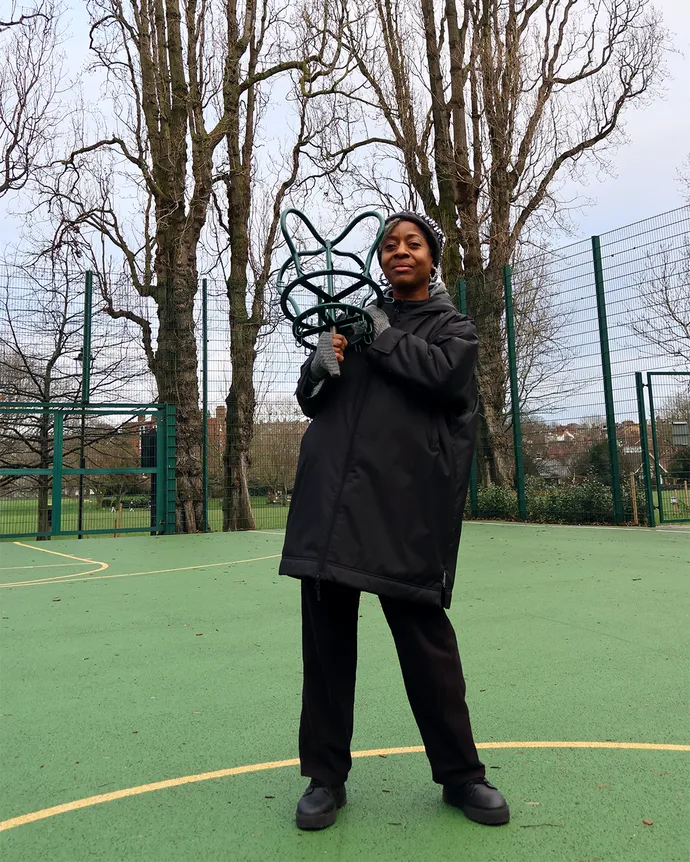 Person in a black coat and gloves holds a green, star-shaped object on a basketball court with tall trees and a fence in the background.