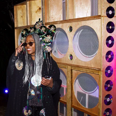 Person in a CD-adorned outfit and ornate hat poses in front of a wooden structure with circular patterns, smiling and holding a phone.